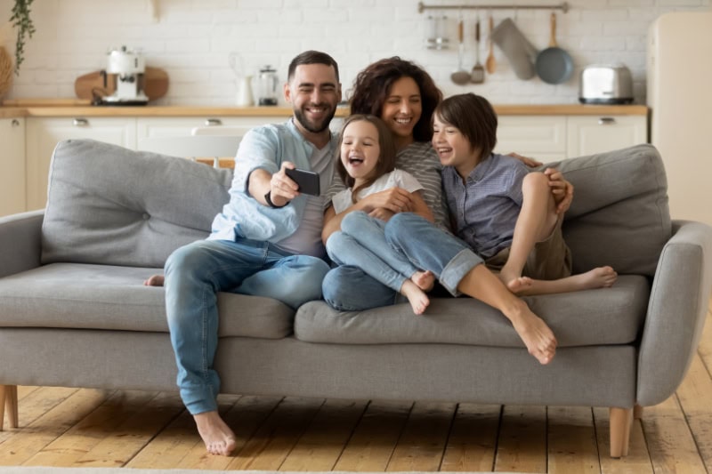 What Are Signs I Need a New Heating and/or Cooling System? Photo of a family of four sitting on a couch laughing and posing for a photo using father's cell phone.